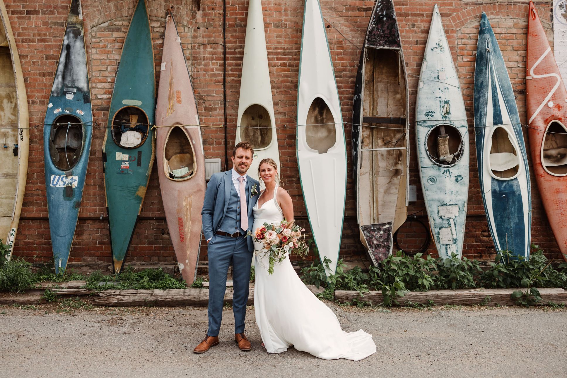 bride and groom standing in front of old kayaks