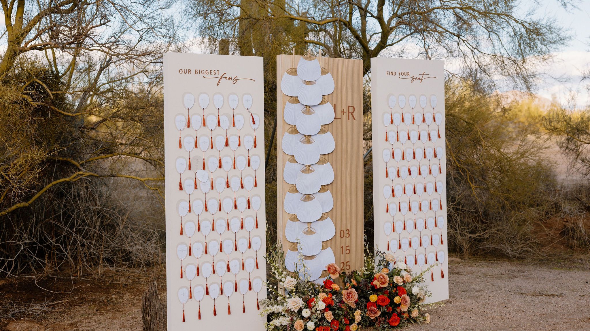 Outdoor wedding escort card display with two guest seating boards, a wood panel with white fans and colorful florals