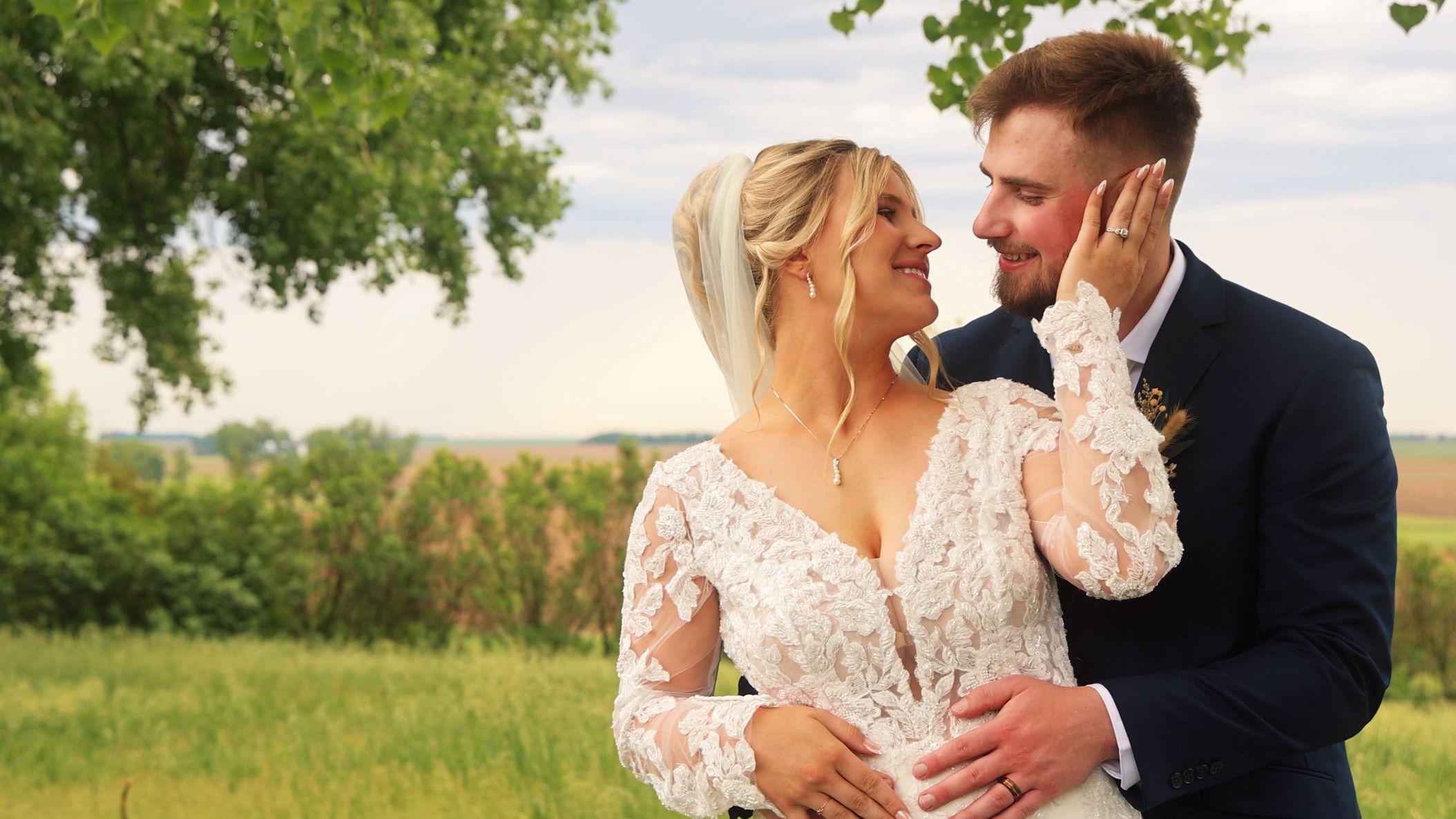 Bride in a long-sleeved lace wedding gown smiles at her groom in a navy suit, touching his cheek as they embrace outdoors.