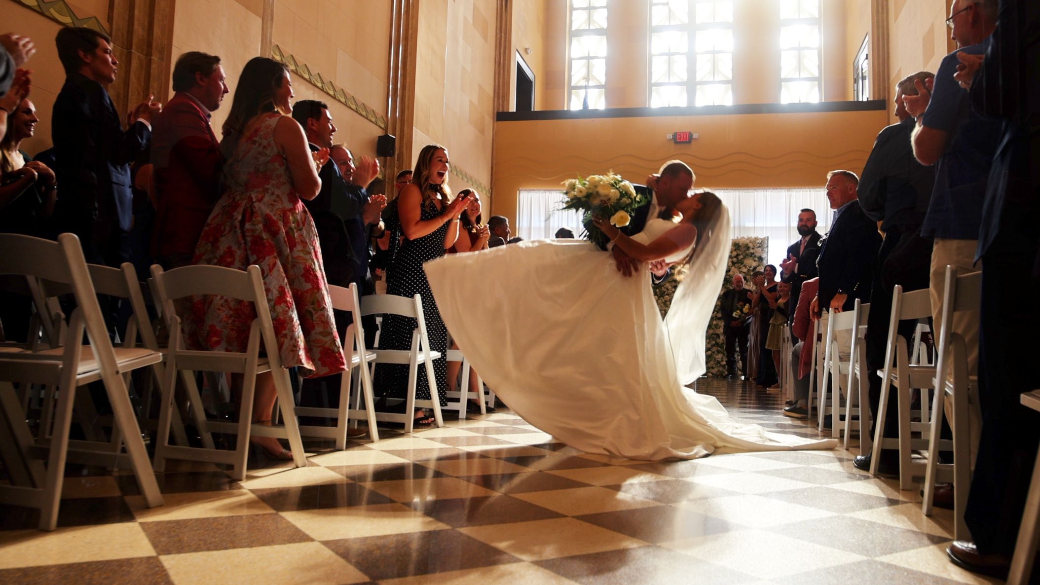Bride and groom share a dramatic dip and kiss in the wedding ceremony aisle as guests stand and applaud on either side.