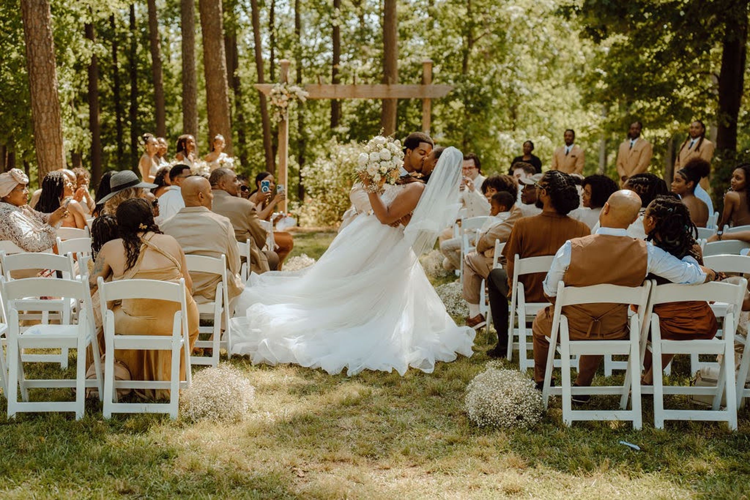 Outdoor wedding ceremony featuring a bride and groom sharing a kiss, surrounded by guests in a lush forest setting, with floral arrangements by Poppy Flowers