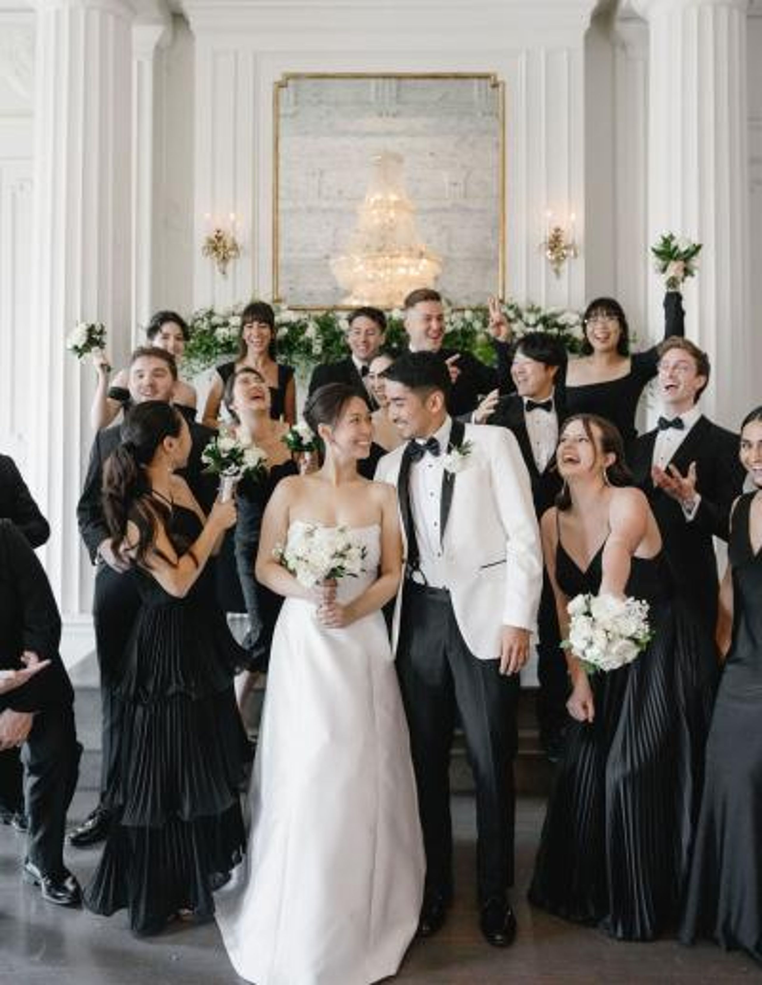 Wedding party featuring an elegant, classic bride and groom surrounded by bridesmaids in black dresses and groomsmen in tuxedos holding white floral bouquets by Poppy Flowers
