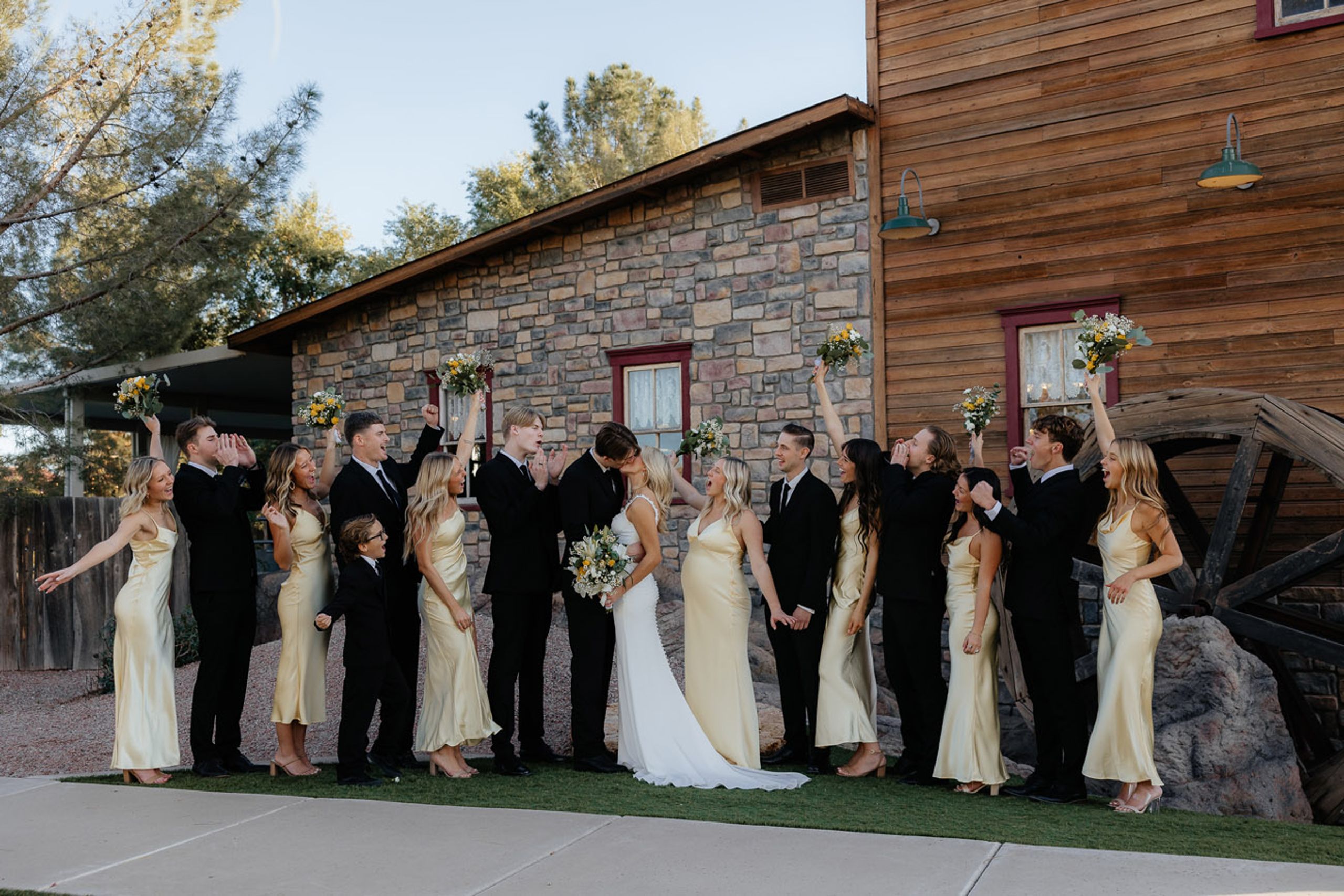 Bride and Groom share a kiss at rustic venue cheered on by their bridesmaids and groomsmen