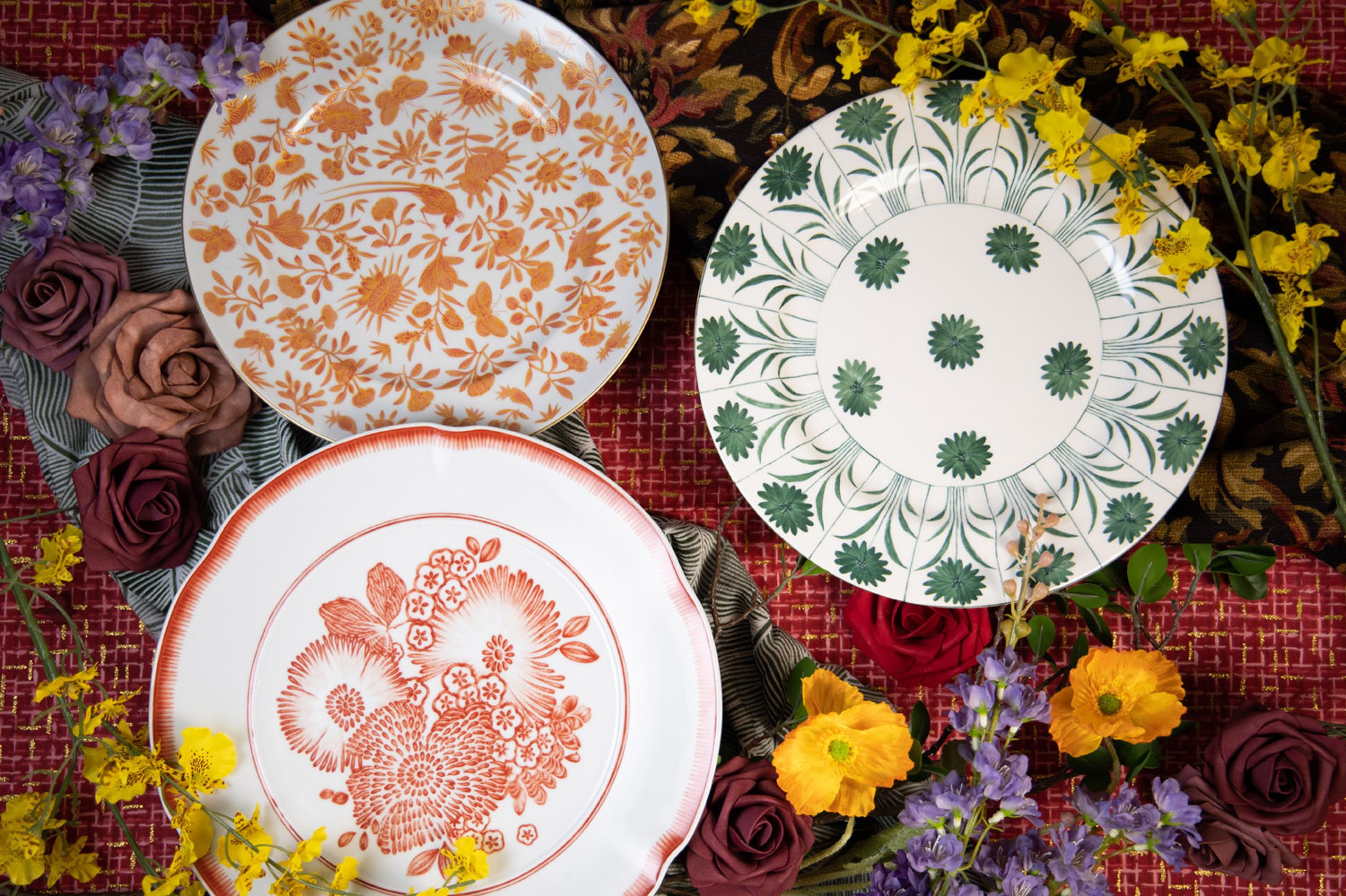 Three designer plates from Maison de Carine—orange Sacred Bird and Butterfly, green Topiary, and coral Oscar’s Coral—styled with roses, poppies, and patterned linens
