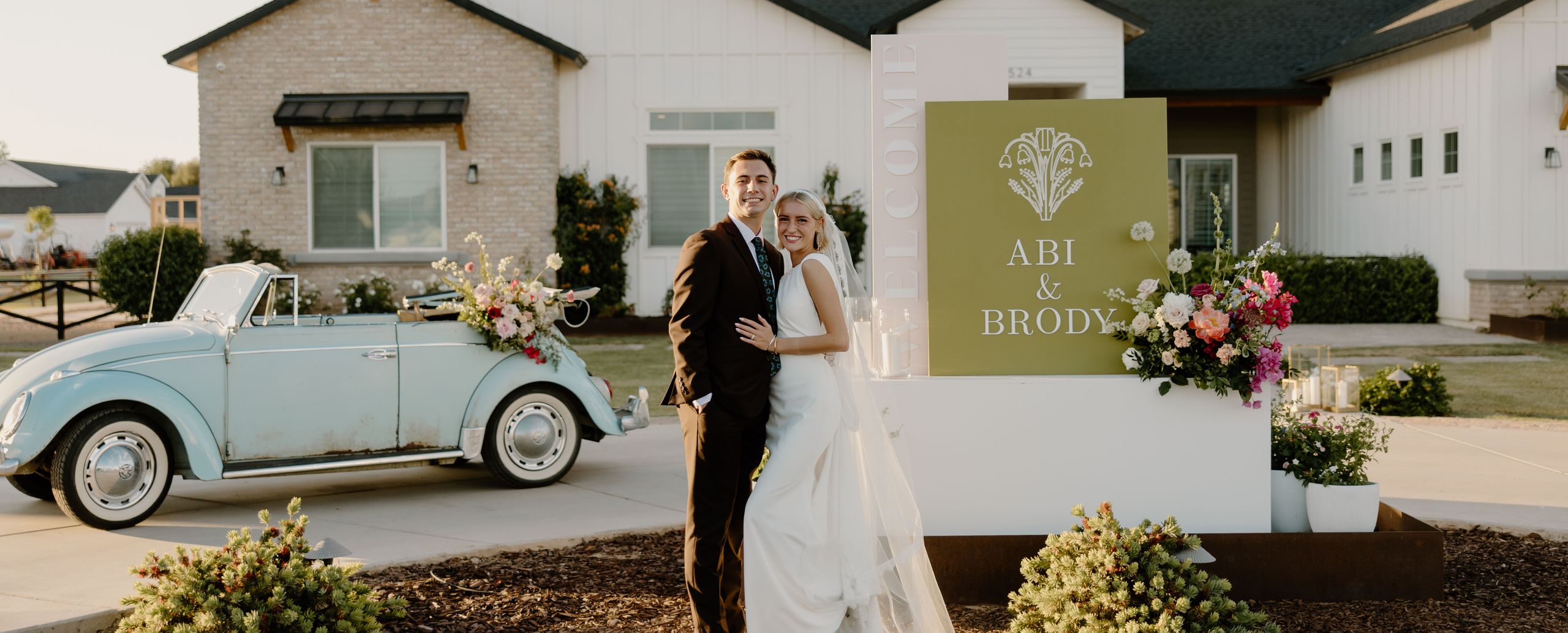 Bride and groom pose in front of groom's house, vintage getaway car and welcome sign