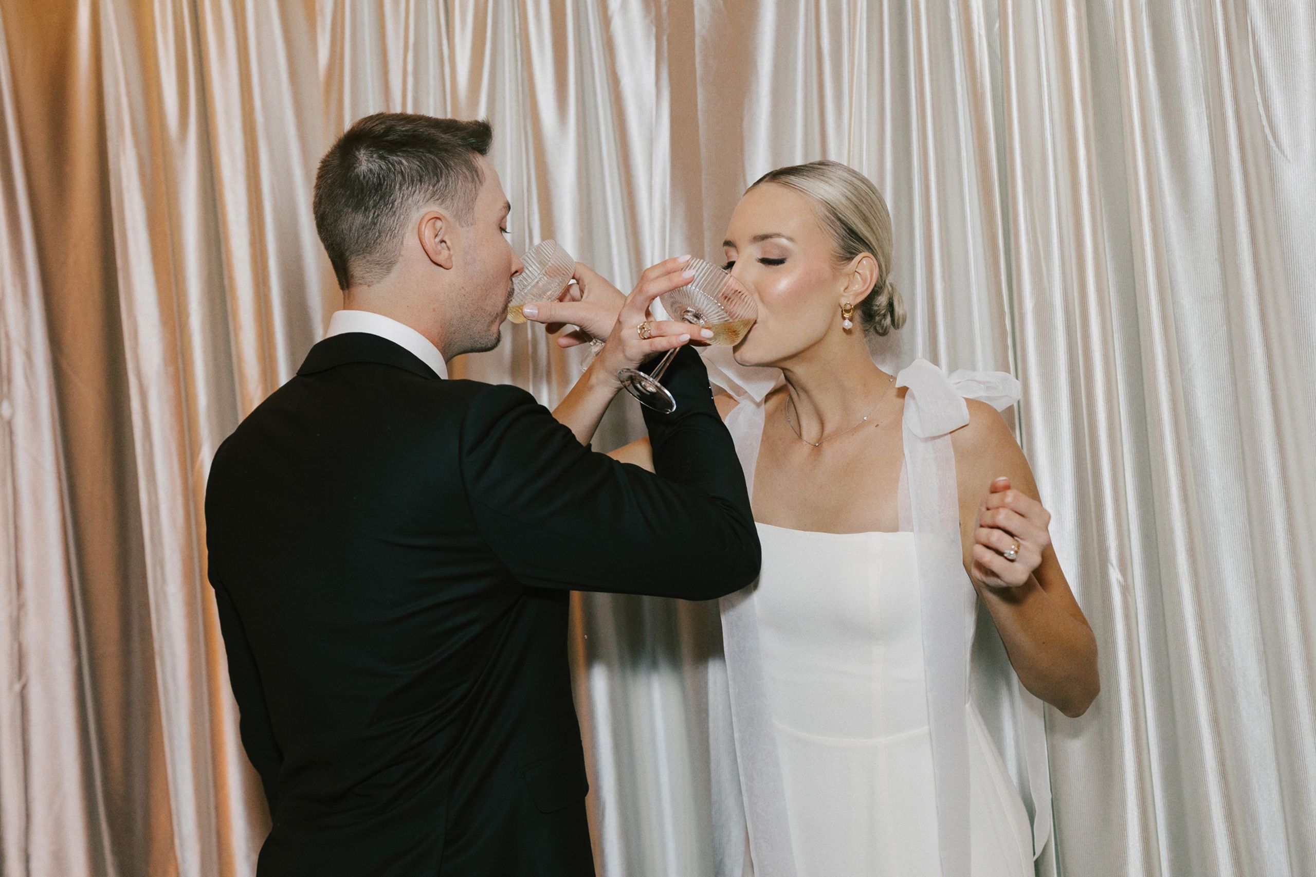 Bride and groom intertwine arms, sipping champagne against a draped backdrop