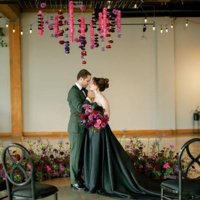Bride in a black wedding dress and groom in a green suit standing beneath hanging pink and purple flowers at their modern indoor winter wedding ceremony