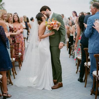 Bride in slip dress, long veil and colorful bouquet, groom in green suit kiss in aisle