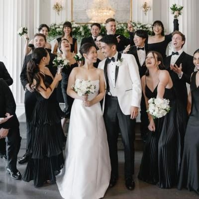 Wedding party featuring an elegant, classic bride and groom surrounded by bridesmaids in black dresses and groomsmen in tuxedos holding white floral bouquets by Poppy Flowers