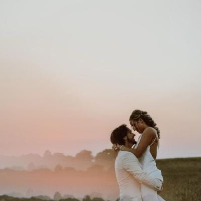 Groom lifts Bride in embrace in a field at sunset
