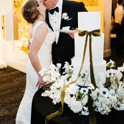 Paige and Johnnie kissing with their cake, surrounded by flowers and a green bow. 
