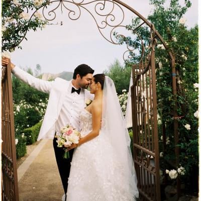 Former Miss California Natasha Alexis Nichelman shares a romantic moment with her groom beneath a vintage wrought-iron gate surrounded by white roses at Tuscan Rose Ranch.