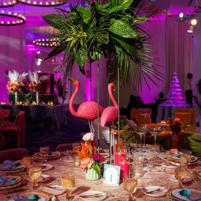 A vibrant dining table at the Curate Gala 2024 features pink flamingo centerpieces, lush tropical greenery, gold flatware, and teal napkins, embodying the Palm Royale theme in the ballroom decor