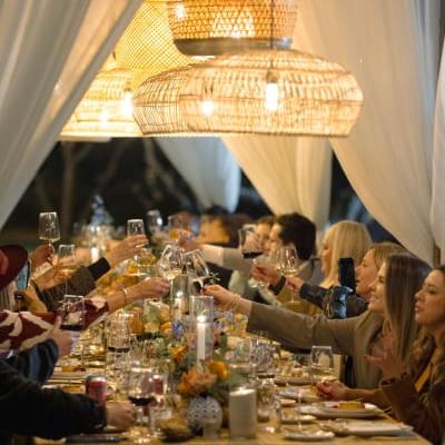 Guests raising wine glasses in a toast under warm, glowing woven chandeliers at a beautifully decorated outdoor dinner table with floral arrangements and candlelight