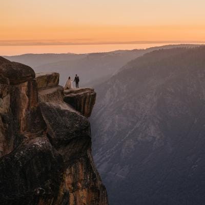 Bride and groom standing on the edge of Taft Point in Yosemite National Park at sunset, overlooking the expansive valley below with stunning cliffs in the background.