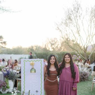 Two stylish women standing next to a modern event welcome sign surrounded by lush purple and green floral arrangements at an outdoor wedding industry gathering.
