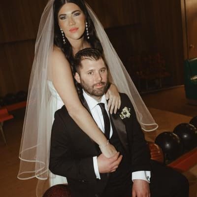 A newlywed couple poses at a vintage bowling alley, exuding confidence and fun. The bride, wearing a classic veil and modern gown, drapes her arms over the groom, who is dressed in a sleek black suit, as bowling balls line the table in front of them.