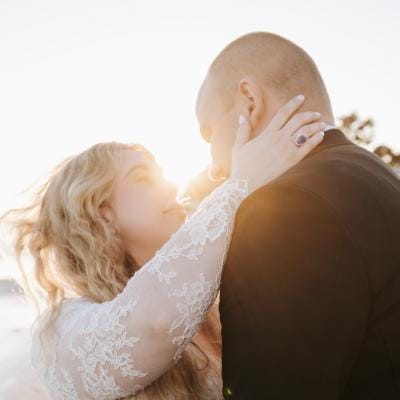 Bride and groom embrace during golden hour on the beach at Rosewood Miramar Beach, bride wearing a lace-sleeved gown and groom in a classic black tuxedo