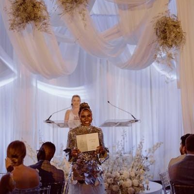 An honoree in a silver sequin top and tiered white tulle skirt holds their award on stage at the Wisconsin Bride Best of 2025 event, framed by cloud-like florals and soft draping decor.