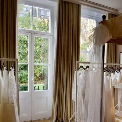 Bridal gowns from E&W Couture displayed on garment racks inside a London suite during the New Archive collection launch, with natural light and garden views in the background.