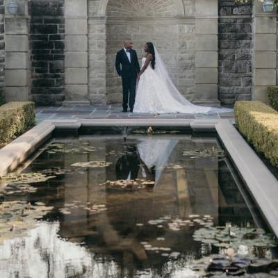 Bride and groom pose by pond, framed by stone architecture at Greystone Mansion