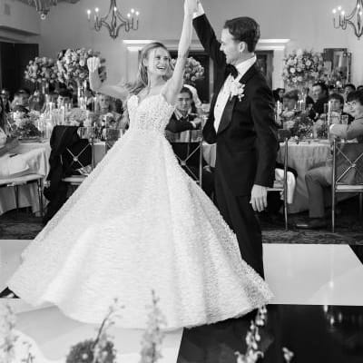 Groom twirls bride on black and white checkered indoor dance floor while their friends and family look on from their tables