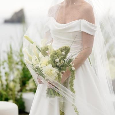 Model poses with a bouquet of lush greenery in an off the shoulder gown and veil with the coast of Crook Point in the background