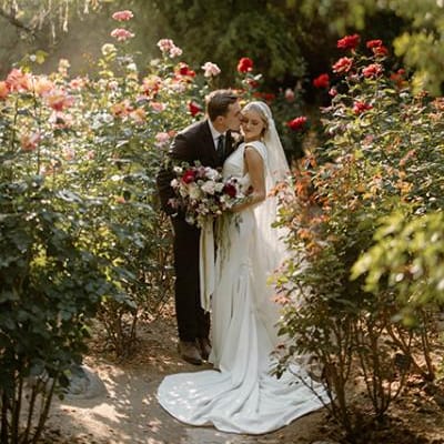 Groom kisses bride on the cheek, surrounded by vibrant flowers and bathed in sunlight