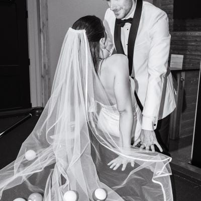 Bride and groom lean against the pool table with pool balls scattered throughout the bride's veil, in black and white