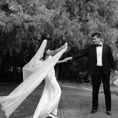 Black and white portrait of bride approaching her groom, her long veil trailing behind her