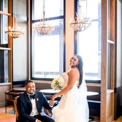 Groom sits and bride stands next to him, posing in the lobby of The Trade Hotel