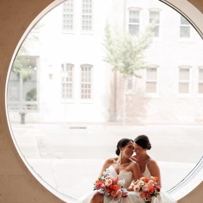 Caroline and Amy cozy up against a large circular window on their wedding day at Overture Center for the Arts