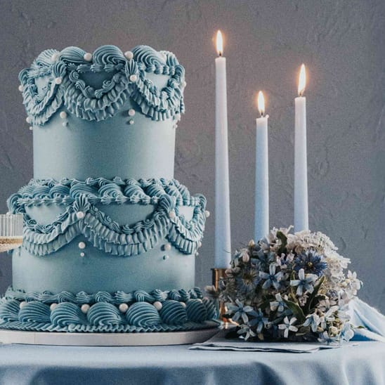 Dusty blue styled scene featuring two-tiered lambeth cake, candles and two glasses of champagne on silky tablecloths