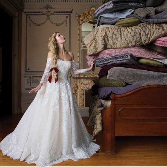 In "Princess and the Pea" style, the bride, in a lacy off-the-shoulder gown, contemplates a bed piled high with mattresses.