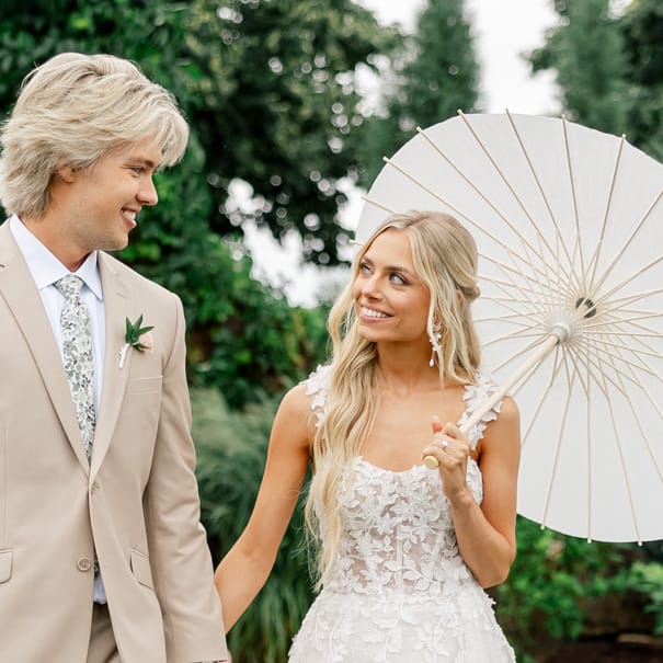 Bride and groom pose in the middle of their outdoor aisle lined by pale pink blooms at Redeemed Farm, bride has a paper umbrella in her hand