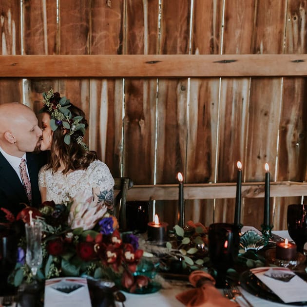 Bride in flower crown with tattoo sleeve and groom sit at a styled table with black tapers burning bright. Shot at The Barn at Wagon Wheel Farm.