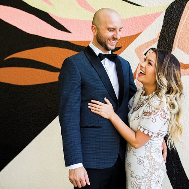 The couple stands together in front of a dusty pink and orange mural at Coopers Hall