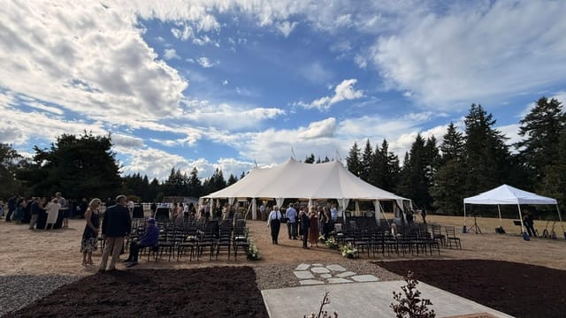 Large white wedding reception tent beside an outdoor ceremony setup beneath a dramatic blue and cloudy sky.
