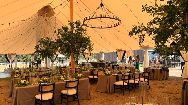 Wedding reception beneath a sailcloth tent with long tables, neutral linens, white floral arrangements, string lights and a circular chandelier.
