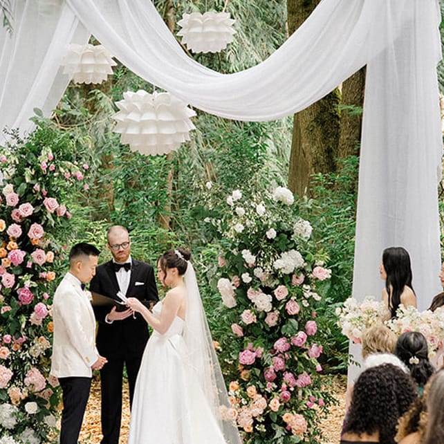 Bride and groom stand at draped outdoor altar for their ceremony, surrounded by their bridal party at Twin Willow Gardens