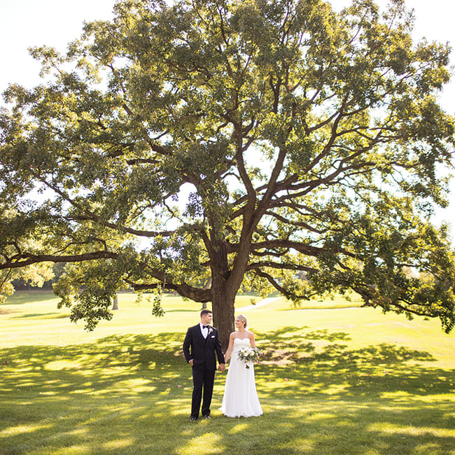 Jake and Paige hold hands in front of a tree during their wedding at the Wayzata Country Club,