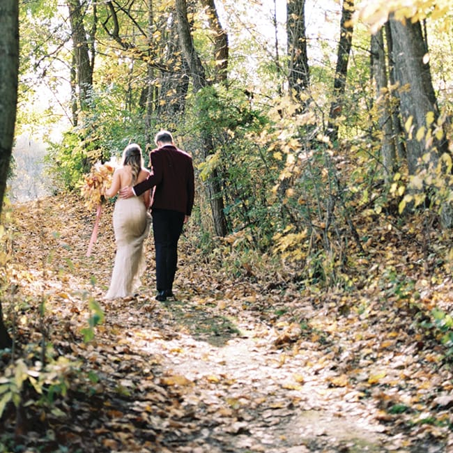 A bride and groom walk a path at the golden hour. His burgundy suit and her neutral wedding dress complement the fall wedding theme.