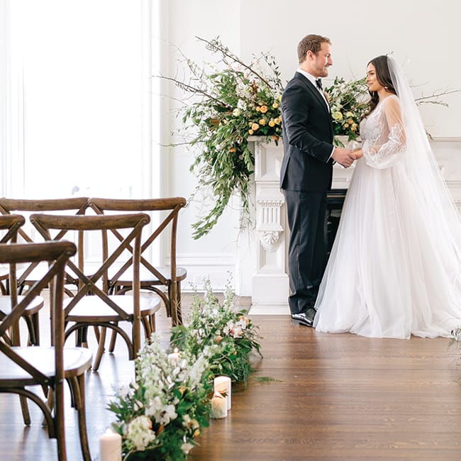 A bride and groom stand in the aisle at the Covenant at Murray Mansion amid their orange, gold and peach wedding flowers.