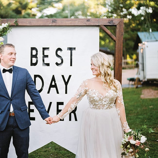 Ashley Zelenka and Evan Bohman hold hands in front of a Best Day Ever sign at their rustic camp wedding.