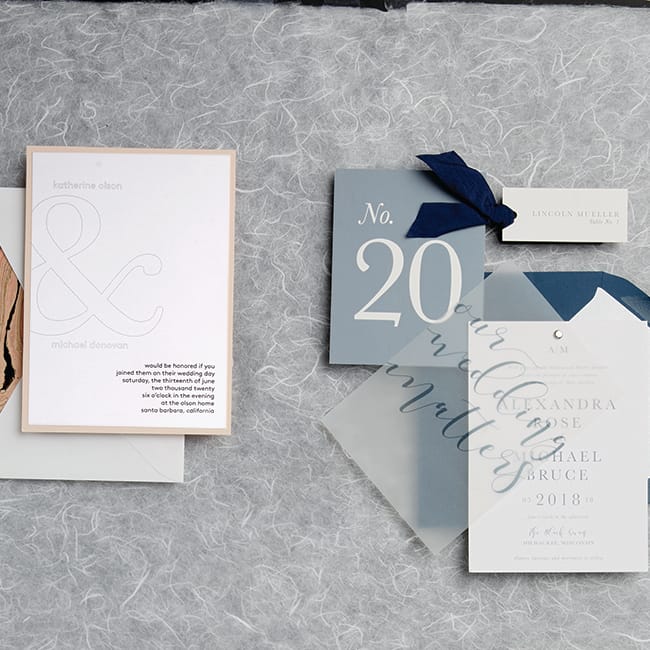 Two wedding invitations, one featuring embossed names and a marbled design and the other featuring a vellum overlay and the wedding hashtag