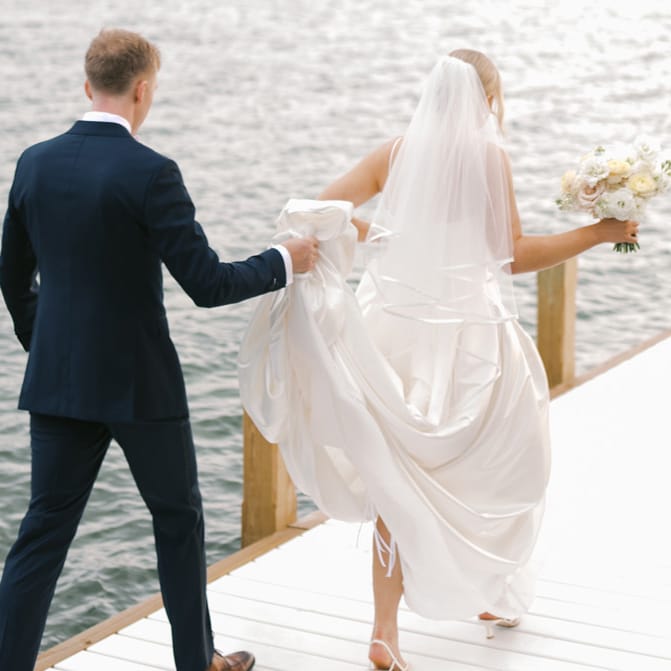 Grace and Lucas walk out on a dock of a lake in their wedding day attire