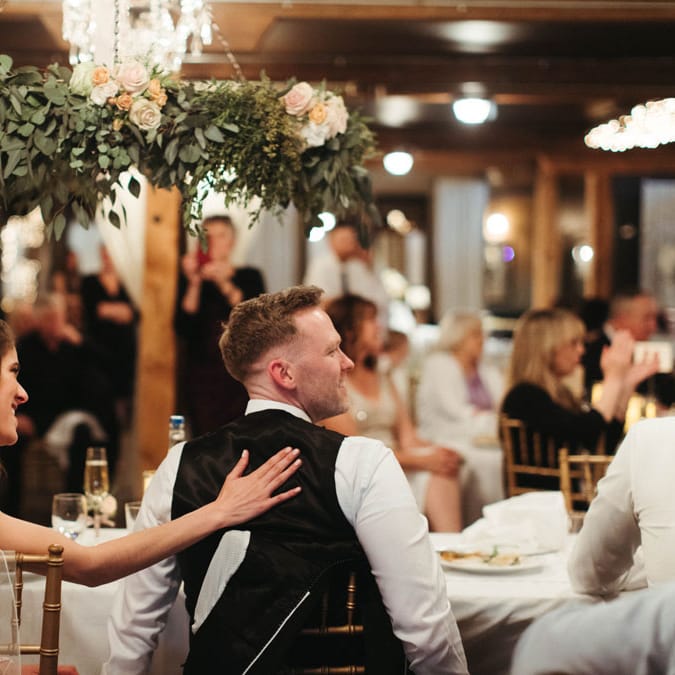 Bride and groom smile at their reception as they sit at their sweetheart table under a custom floral installation by Belles + Whistles