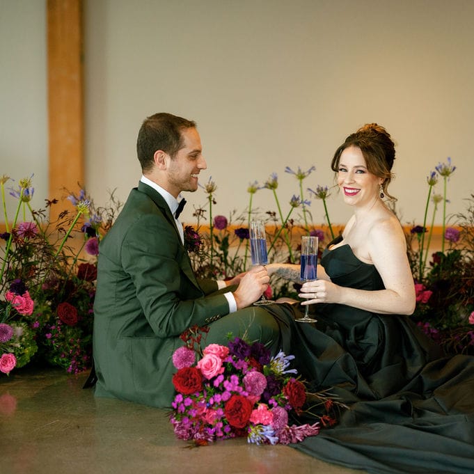 Bride and groom sitting on the floor surrounded by bold winter florals, sharing a drink during their colorful indoor wedding celebration