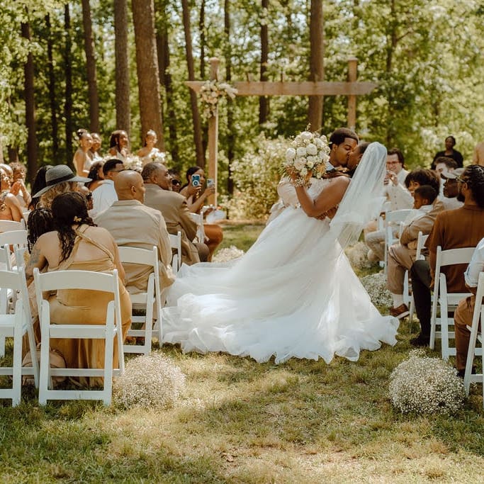 Outdoor wedding ceremony featuring a bride and groom sharing a kiss, surrounded by guests in a lush forest setting, with floral arrangements by Poppy Flowers