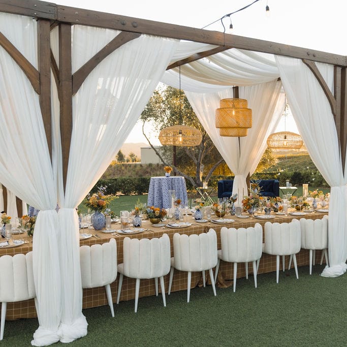 Elegant outdoor dining setup with draped pergola, modern white chairs, and a long table adorned with floral centerpieces and patterned vases under woven chandeliers at sunset